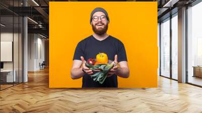 Charming young hipster man with beard holding raw vegetables over yellow background. Wall mural