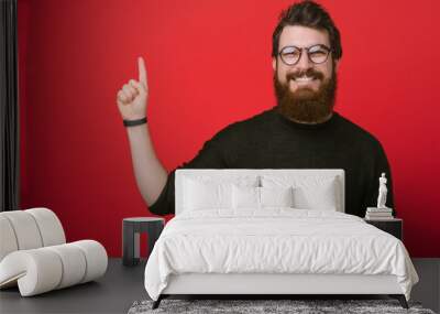 Cheerful bearded man in glasses, holding a smartphone and pointing finger at copy space Wall mural