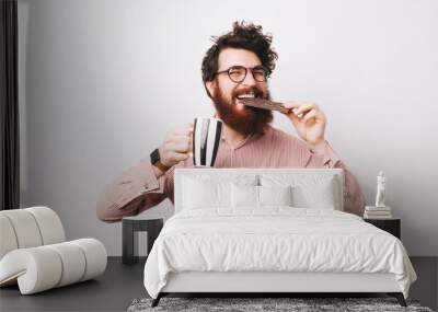 Cheerful happy bearded man in causal eating bar of chocolate and holding cup of coffee Wall mural