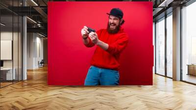 Excited man with beard, playing in mobile phone, over red background Wall mural