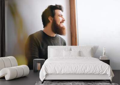 Handsome cheerful bearded man sitting near window, thinking and looking forward while using laptop Wall mural