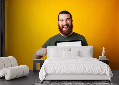 happy bearded hipster pointing at his blank white screen of the notebook on yellow background. Wall mural