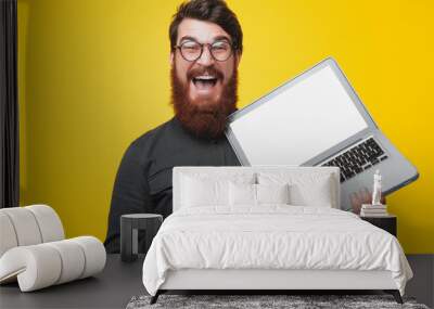 Happy bearded man in glasses holding and pointintg at laptop while looking at camera Wall mural