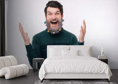 Photo of cheerful surprised young man in green pulover and decorated christmas beard gesturing over white background Wall mural