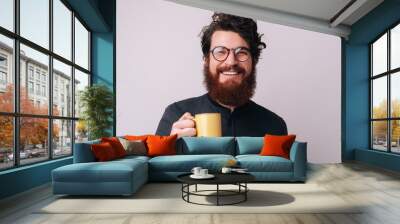 Photo of some cheerful bearded guy wearing glasses, loooking at camera and holding a mug with coffee, over isolated background Wall mural