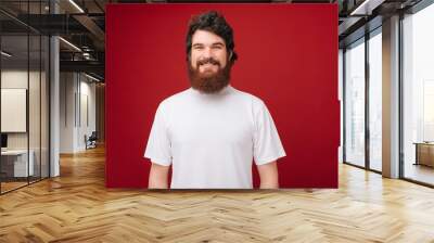 Portait of cheerful bearded guy in white tshirt smiling and looking at camera over red background Wall mural