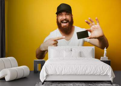 Portrait of a cheerful bearded man pointing finger at credit card isolated over yellow background Wall mural