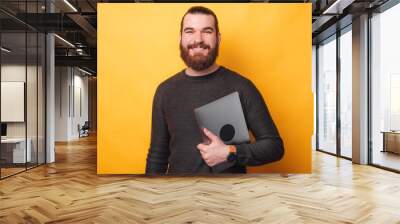 Portrait of handsome young man with beard holding laptop and looking at camera. Wall mural