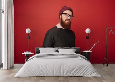 Serious and confident bearded man posing in studio with red background. Handsome man looking at camera, holding notebook. Wall mural