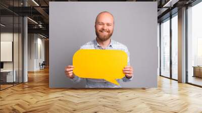 Smiling handsome man standing over grey background and holding yellow speech bubble with copyspace. Wall mural