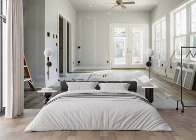 An interior of a house being renovated, showcasing new wooden flooring, a ladder, wall patches, and a ceiling fan. Bright windows let in natural light into the room. Wall mural