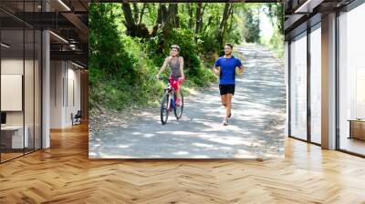 young woman riding bicycle beside a young man running in countryside in summer Wall mural