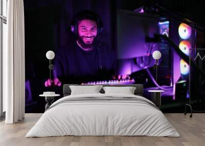 A young man plays a computer game in a room with colorful neon lights. raising fists merrily with satisfaction in the black studio A man wearing headphones intends to play a game online. Wall mural