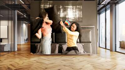 Happy mixed-race lesbian couple having fun during a pillow fight on a bed mattress. Two cheerful teenage lesbians laughing and playing funny active games in the bedroom, LGBTQIA pride month concept. Wall mural