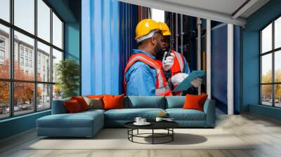 Two attractive black workers in safety helmets and uniforms work at the container port terminal. Two male engineers are checking the products in the cargo container before being exported to the port. Wall mural