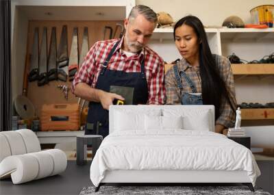 White senior man carpenter doing his work with Asian co worker woman, craft work at furniture factory and copy spcae. Wall mural