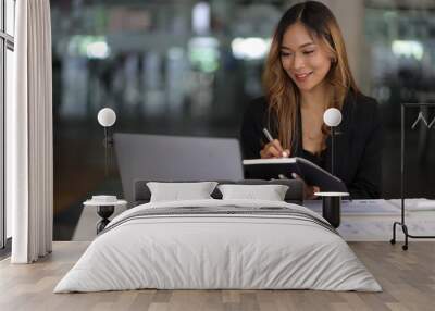 Portrait of young asian businesswoman smiling thinking and writing on notebook while sitting at office desk in modern office. Wall mural