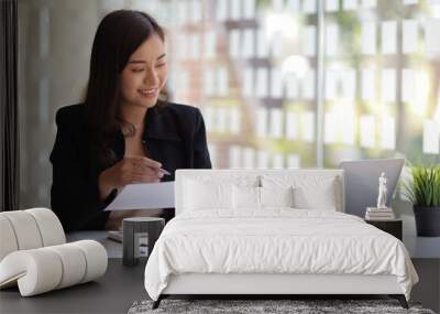 Smiling Asian businesswoman working on laptop and reading documents on desk in office. Wall mural