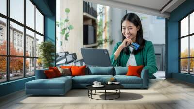 A woman is sitting at a desk with a laptop and a credit card in her hand Wall mural