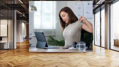 A woman is sitting at a desk with a laptop and a tablet. She is stretching her arms and legs, possibly taking a break from work. Concept of relaxation and self-care Wall mural