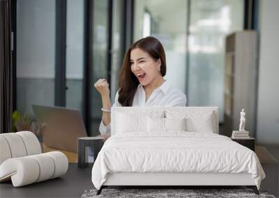 Joyful businesswoman with a victorious gesture, celebrating success while working on her laptop in a sunny office space. Wall mural