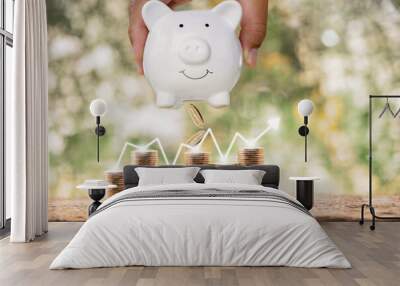 Businessman hand hold the piggy bank put on the stack gold coins with arrow graph chart on transparent screen with growing value ​in the public park, Saving money for future investment plan concept. Wall mural