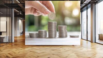 Close-up of a hand stacking coins in ascending order symbolizing financial growth, investment, and savings on a blurred natural background. Wall mural