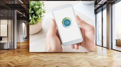 Close-up of hands holding a smartphone displaying a fitness app with plant and desk in the background. Wall mural