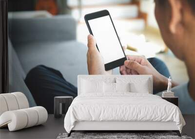 Cropped shot of a young man using mobile phone with empty screen and sitting on sofa in living room at home. Wall mural