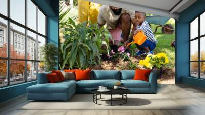 African american father looking at cute son watering fresh flowers with can in backyard Wall mural