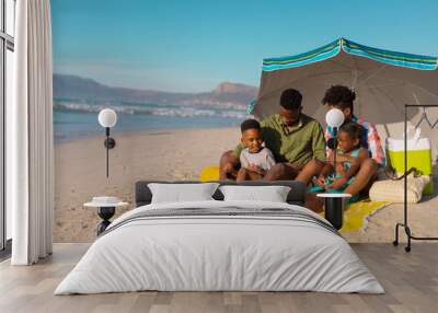 African american young parents with children sitting under umbrella on blanket at beach against sky Wall mural