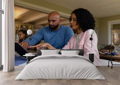 Biracial parents using laptop, looking at bills and talking at table with daughter in background Wall mural