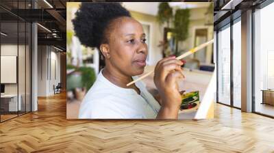 Focused happy senior african american woman holding paint palette painting canvas on easel at home Wall mural