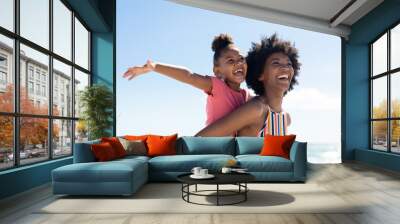 Happy african american woman giving piggyback to daughter with arms outstretched against sky Wall mural