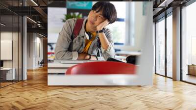 In school, asian teenage boy sitting at desk looking bored with headphones around neck Wall mural