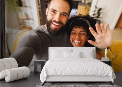 Portrait of happy biracial bearded man waving hand and taking selfie with girlfriend on sofa Wall mural