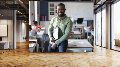 Portrait of smiling african american young male teacher holding file while sitting at desk in class Wall mural