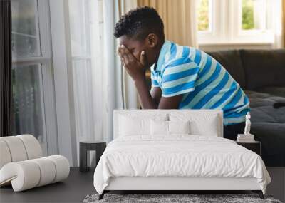 Sad african american boy covering his face and sitting at window in living room Wall mural