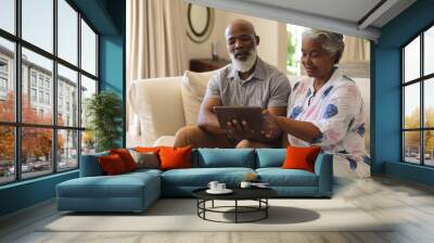 Senior african american couple sitting on sofa using tablet and smiling Wall mural