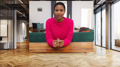 Serious african american young woman looking at camera while sitting at table in living room Wall mural