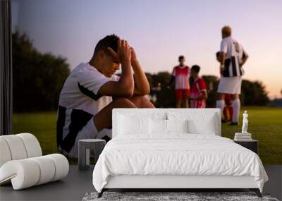 Side view of caucasian sad male player with head in hands and ball sitting on field at sunset Wall mural