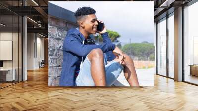 Sitting outdoors, teenage boy talking on smartphone, smiling and enjoying conversation Wall mural