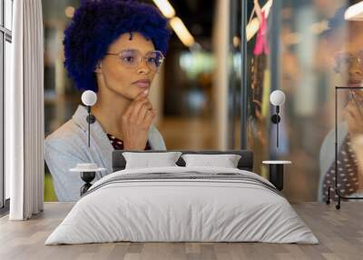 Thoughtful biracial casual businesswoman with blue afro brainstorming at glass wall in office Wall mural