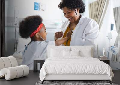 African american female doctor playing with girl patient using stethoscope at hospital Wall mural