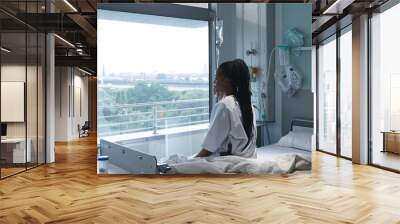 African american female patient sitting on bed and looking through window in hospital room Wall mural