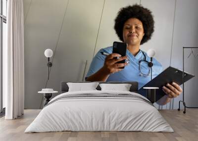 African american mid adult female nurse using smart phone while holding clipboard in hospital Wall mural