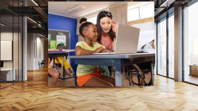 Asian female teacher assisting african american schoolgirl in using laptop at desk in classroom Wall mural