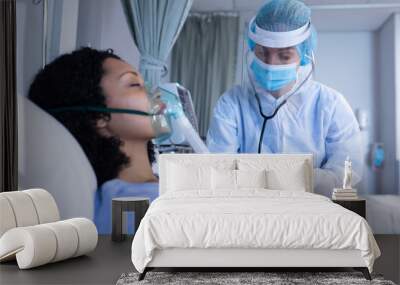 Caucasian doctor in ppe suit examining with stethoscope female patient with oxygen ventilator Wall mural