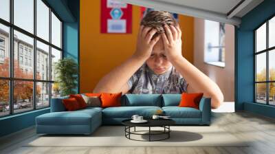 Confused caucasian schoolboy sitting at desk in classroom with books, holding head in hands Wall mural