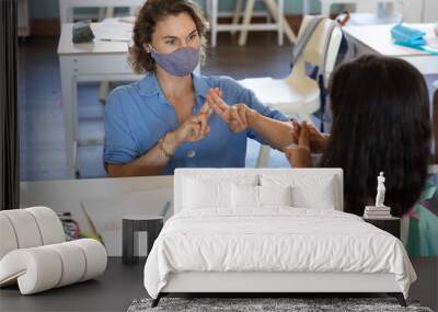 Female teacher and girl wearing face masks talking to each other through sign language in class Wall mural